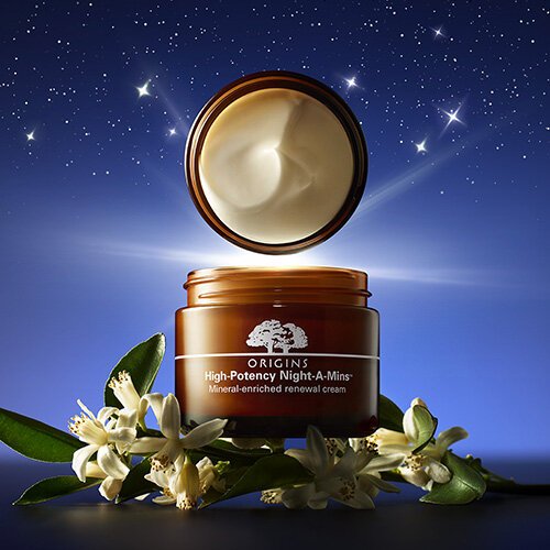 Open jar of Origins High-Potency Night-A-Mins renewal cream, with cream visible inside, surrounded by white flowers and green leaves, against a starry night sky background.