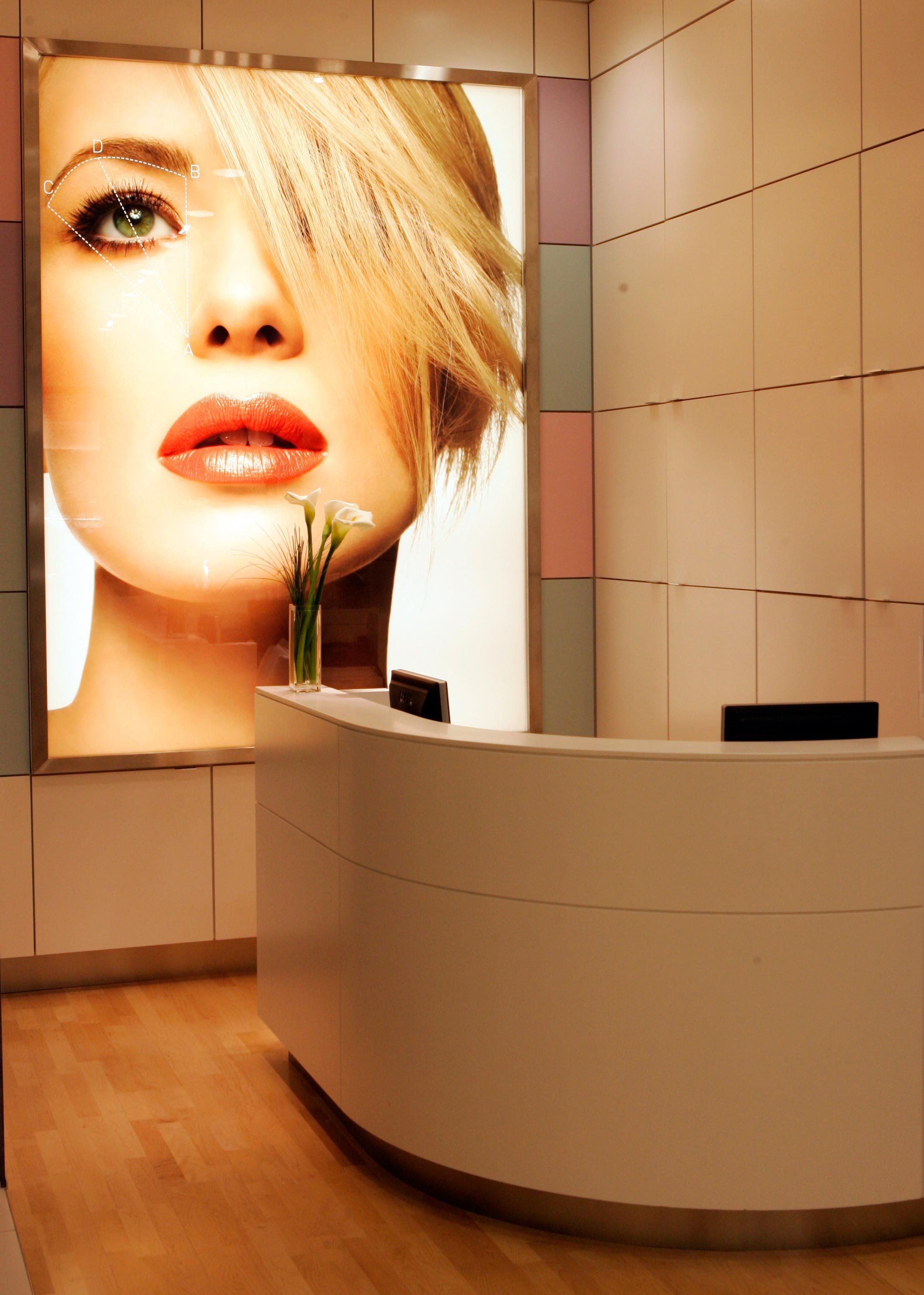 Reception desk in front of a large illuminated advertisement featuring a woman's face with makeup, short blond hair, and green eyes, in a modern interior space.