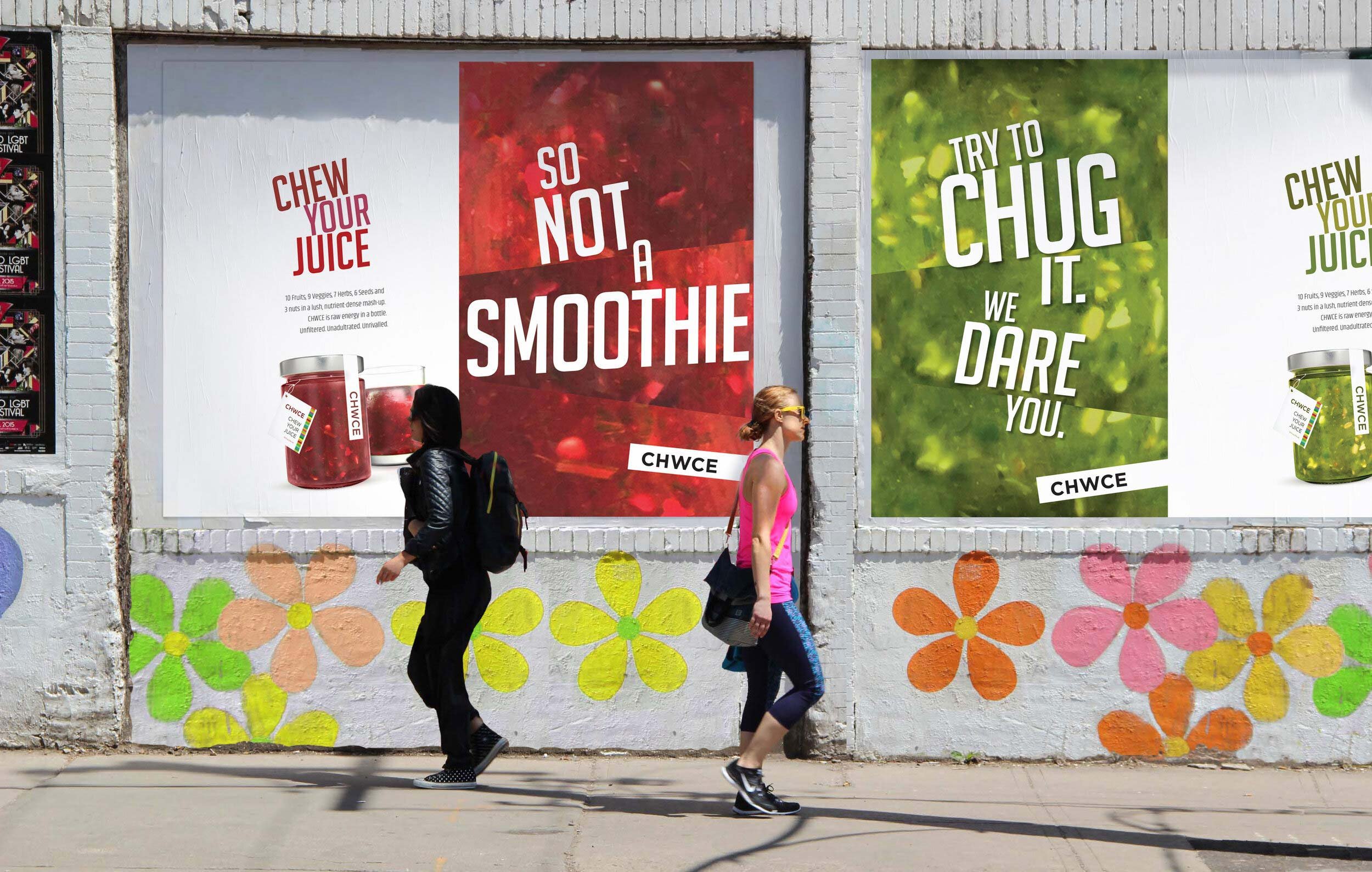 Street scene with two women walking past colorful posters on a white brick wall. The posters promote fruit juice and smoothies, with phrases like 'Chew your juice,' 'So not a smoothie,' and 'Try to chug it. We dare you.' The wall is decorated with pa