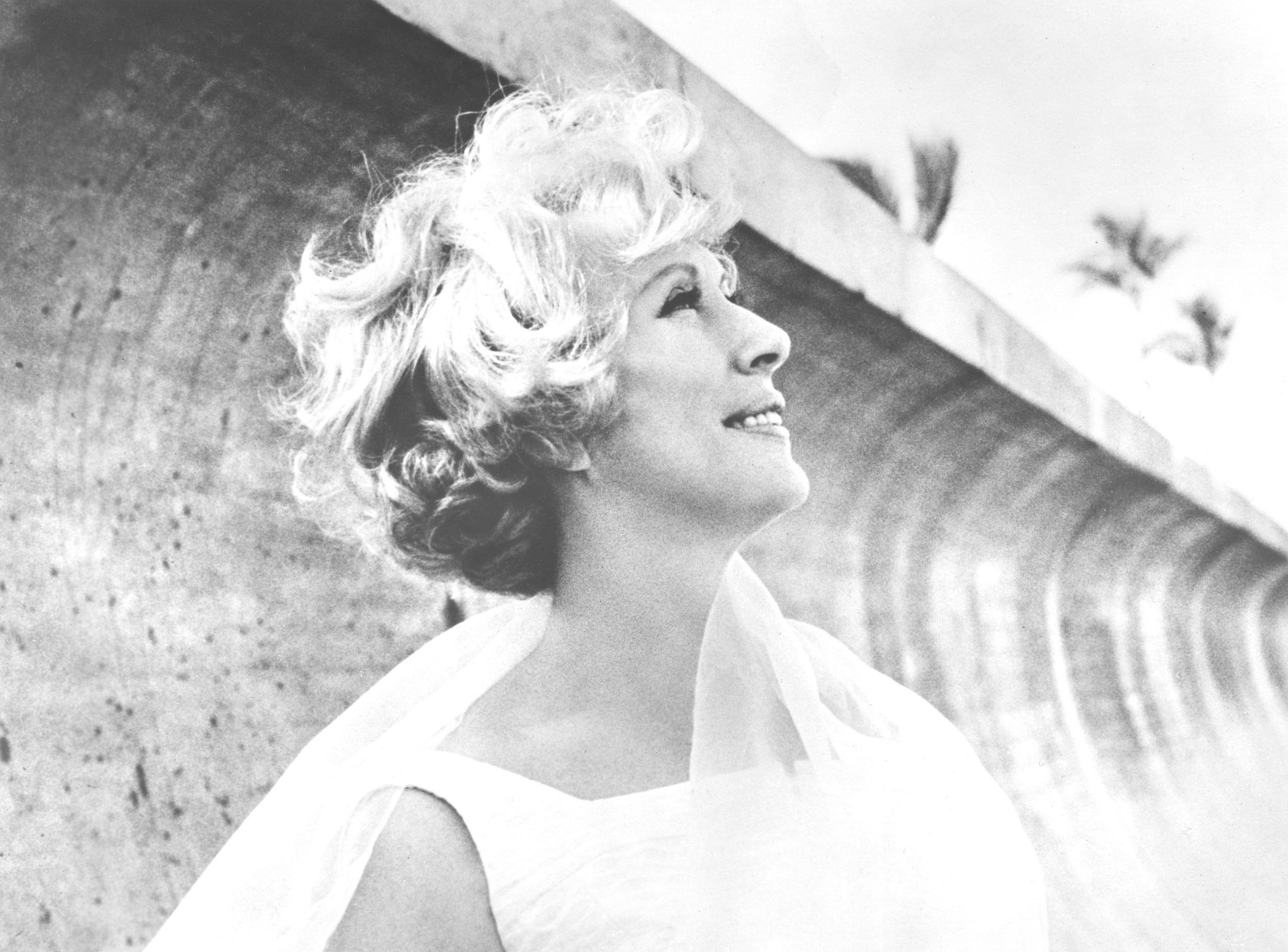 A woman with short, curly blonde hair smiling and looking upward, standing outdoors near a curved concrete structure with palm trees in the background.