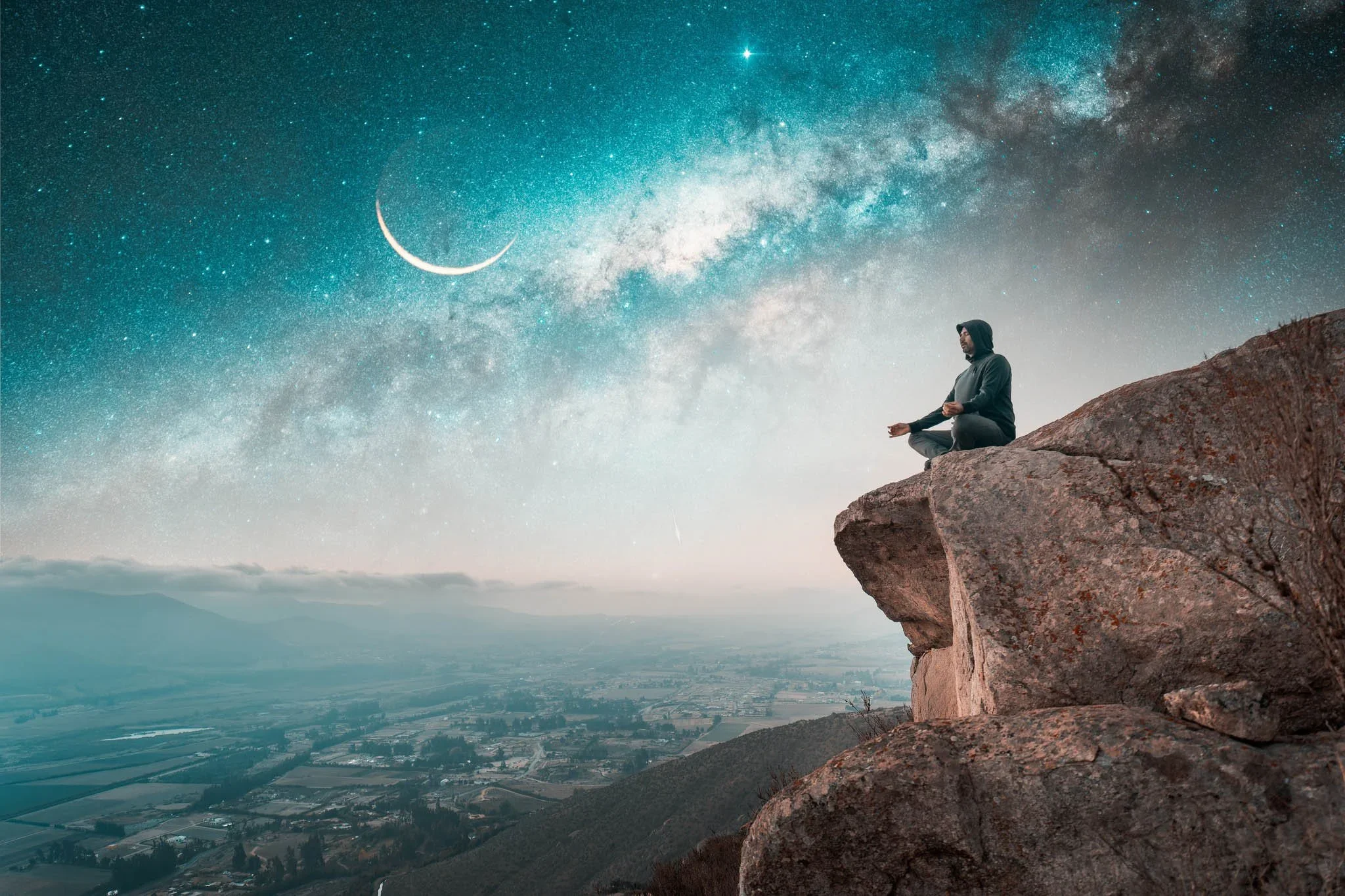 A person sitting cross-legged on a rocky ledge at night, practicing meditation, with a view of the starry sky, moon, and Milky Way galaxy in the background.