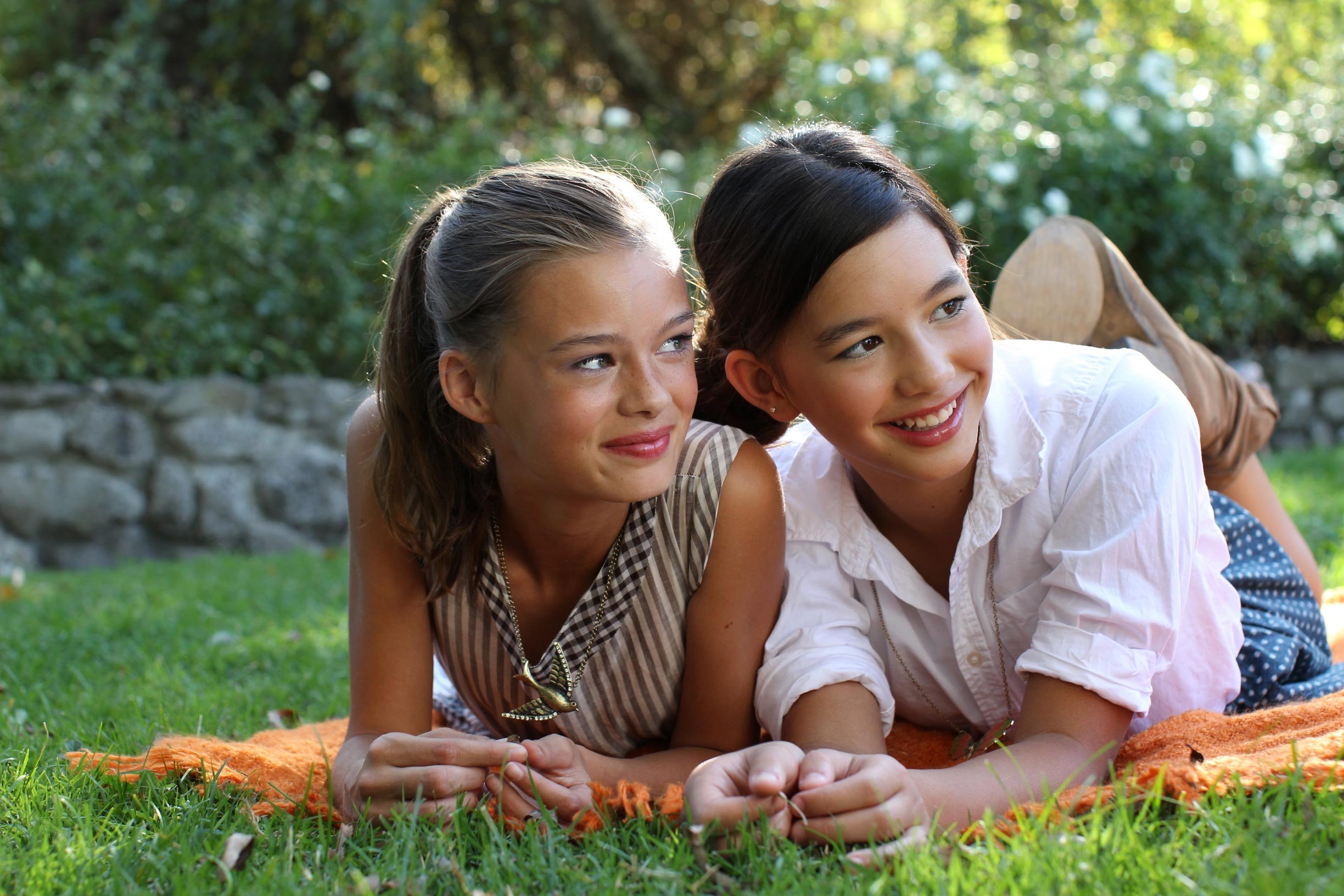 Two young girls lying on a grassy lawn outdoors, smiling and relaxing in the sunlight.