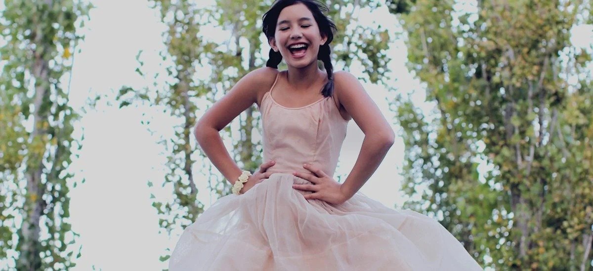 A girl in a light pink dress smiling and sitting outdoors among trees.