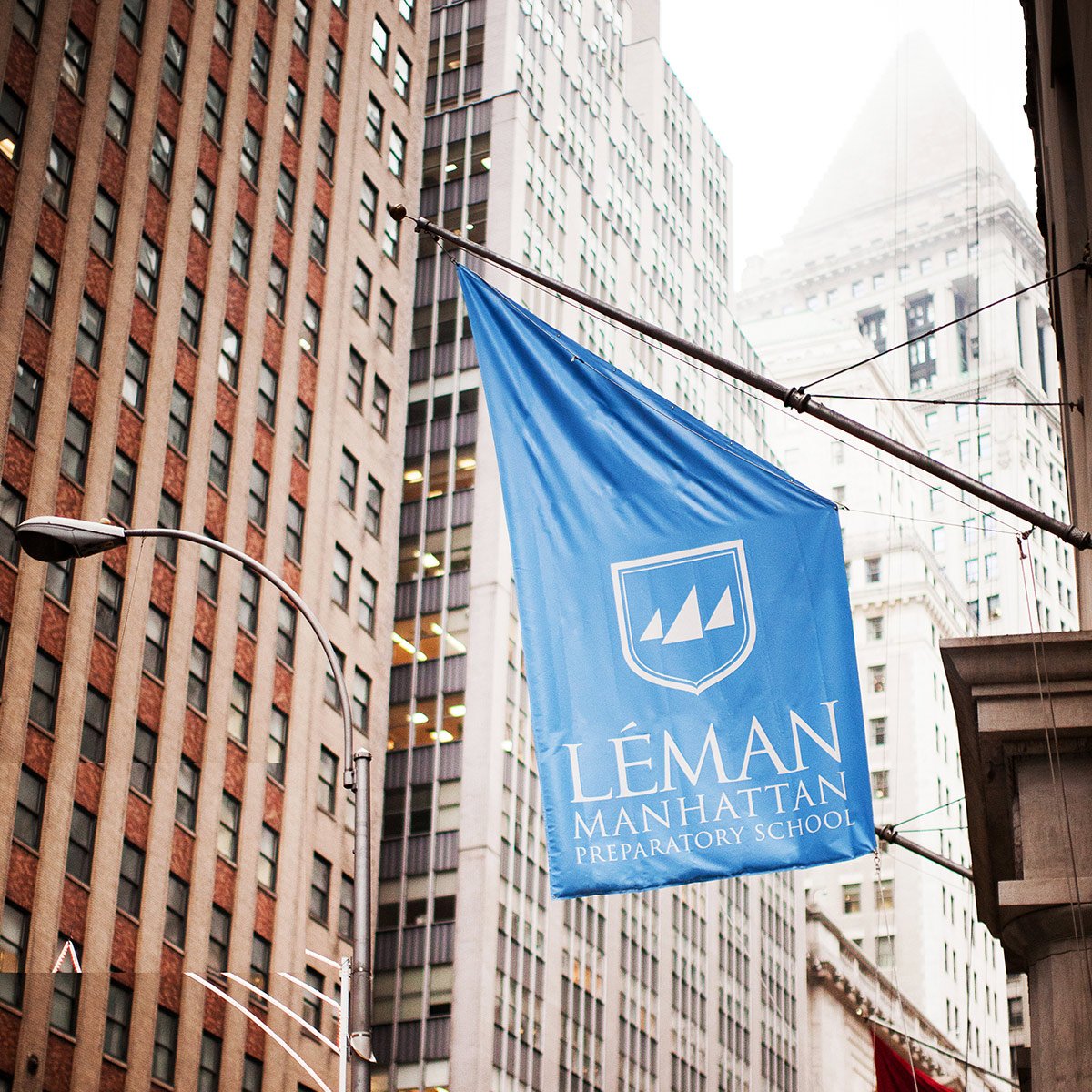 Blue flag with the logo and name of Léman Manhattan Preparatory School hanging between tall urban buildings in Manhattan.