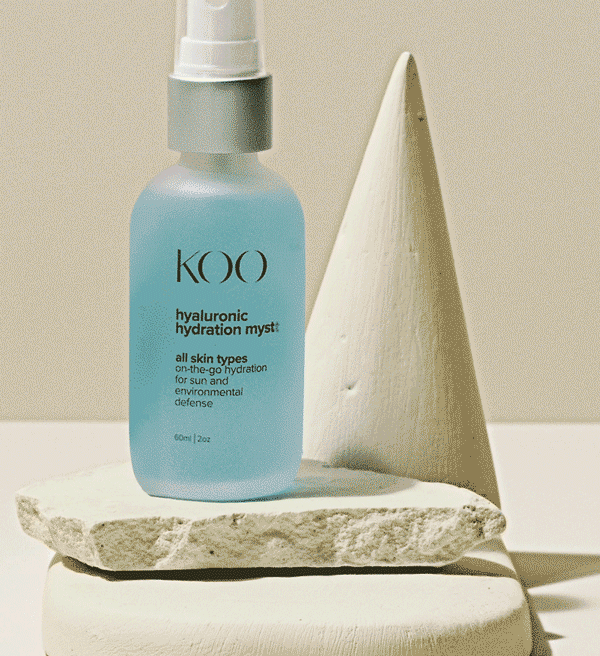 A light blue bottle of KOI hyaluronic hydration mist on a textured white stone, with a white cone-shaped object next to it.