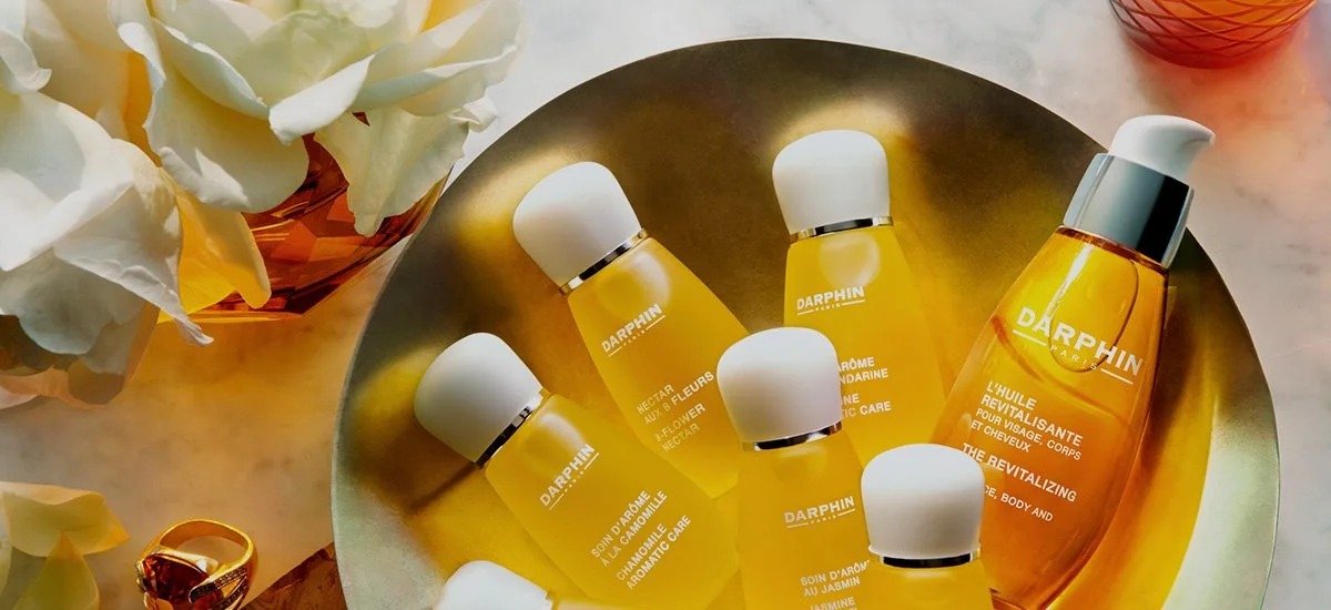 Collection of yellow cosmetic bottles on a silver tray, with a bottle of orange oil, surrounded by white flowers and a gold ring.