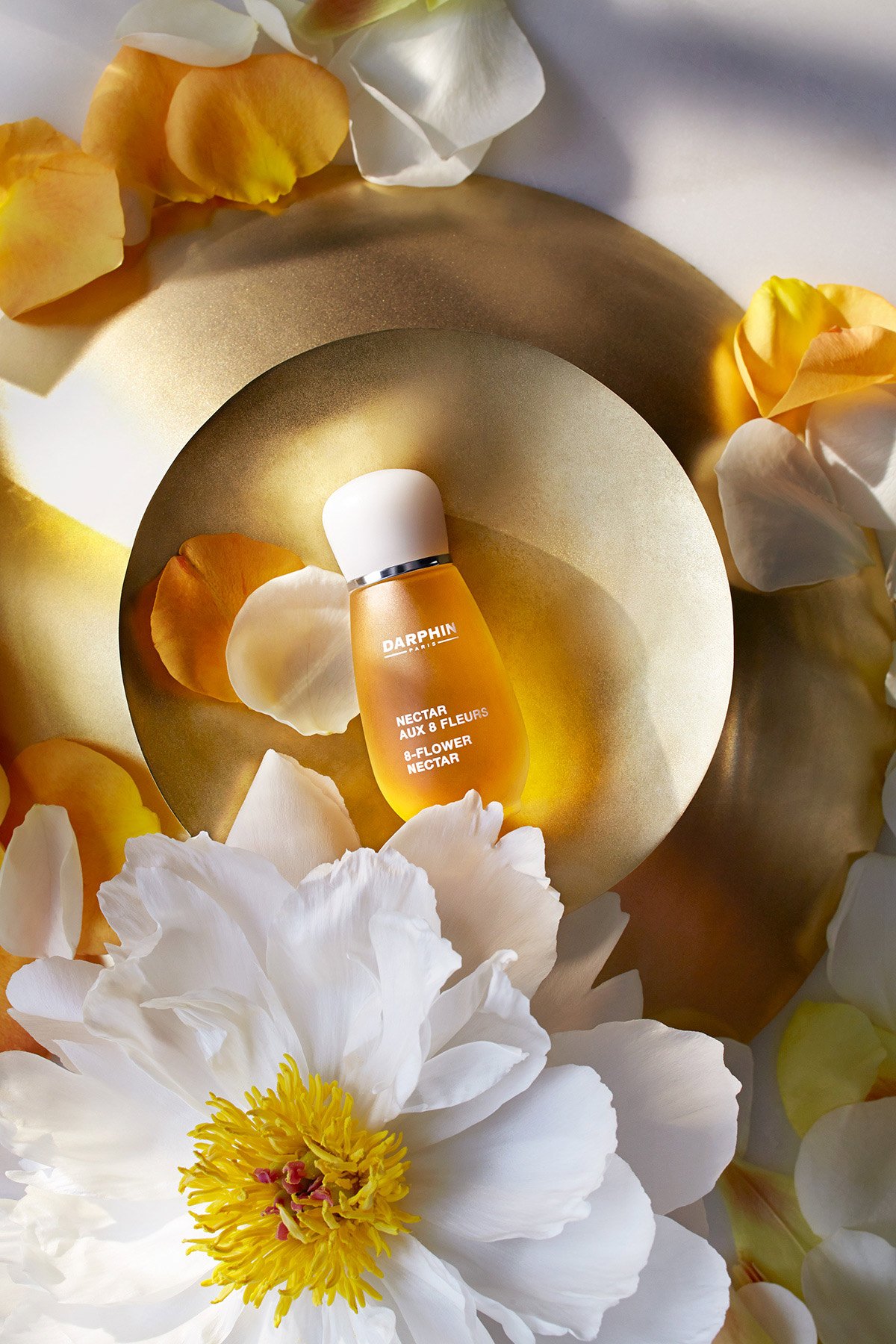 A small golden bottle labeled Darphin Nectar Aux Fleurs 9-Flower Nectar, surrounded by white and yellow flower petals on a gold and white surface.