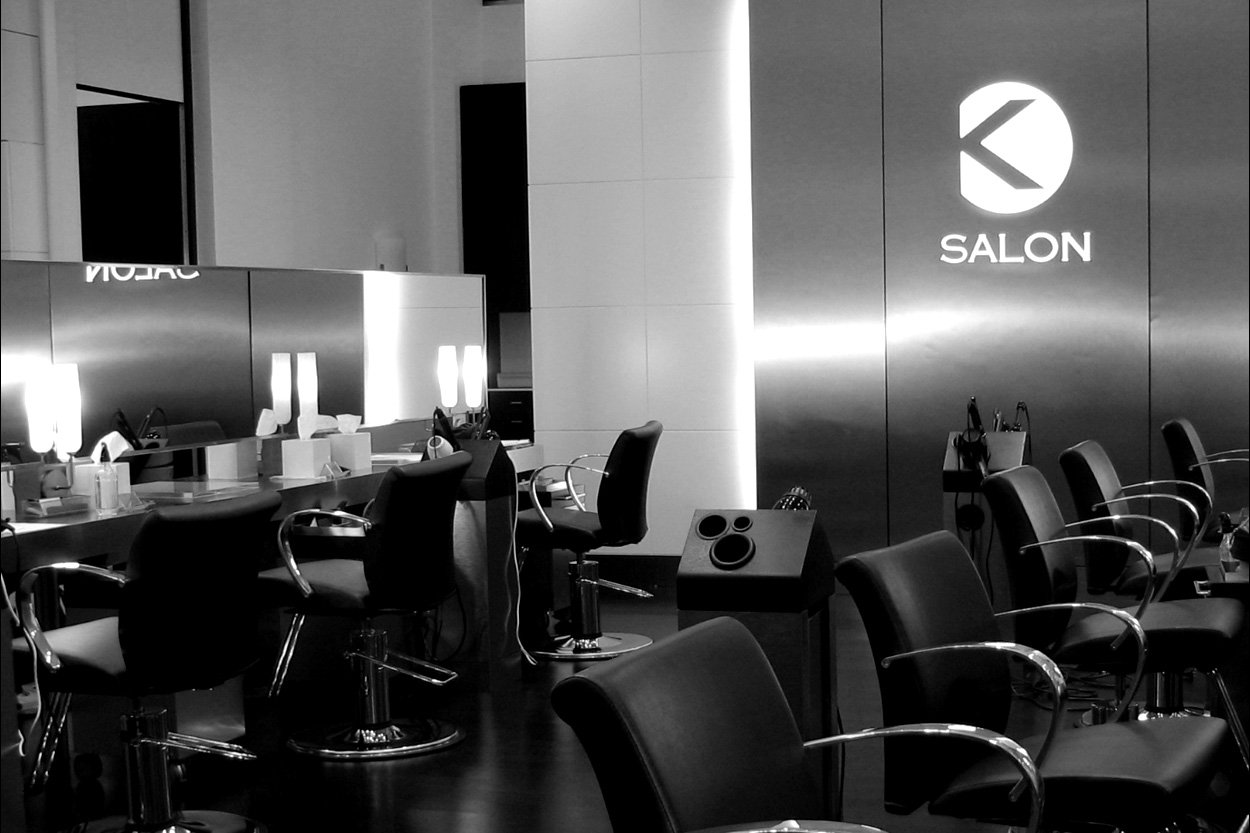 Empty salon with black chairs and mirrors, illuminated by lamps, with a sign that says 'SALON' and a logo on the wall.