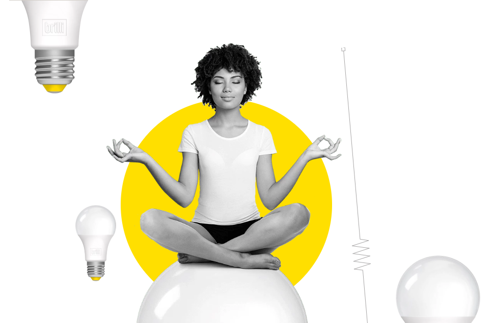 A woman sitting cross-legged in a meditative pose on a large white ball with yellow circle background, surrounded by floating light bulbs and a light bulb pendant, with a large light bulb towards the top left.