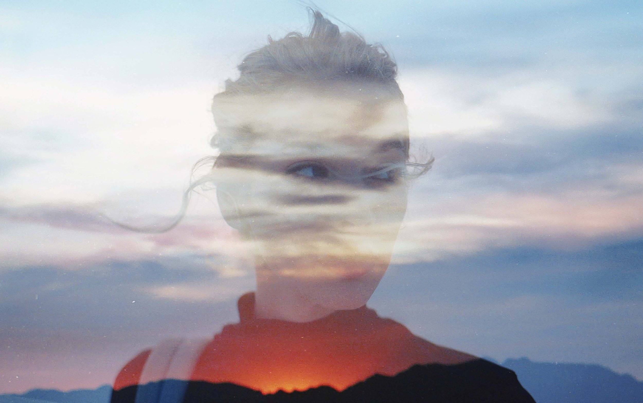 A double exposure photograph showing a woman's face and a sunset sky with clouds, mountain silhouettes, and the horizon.